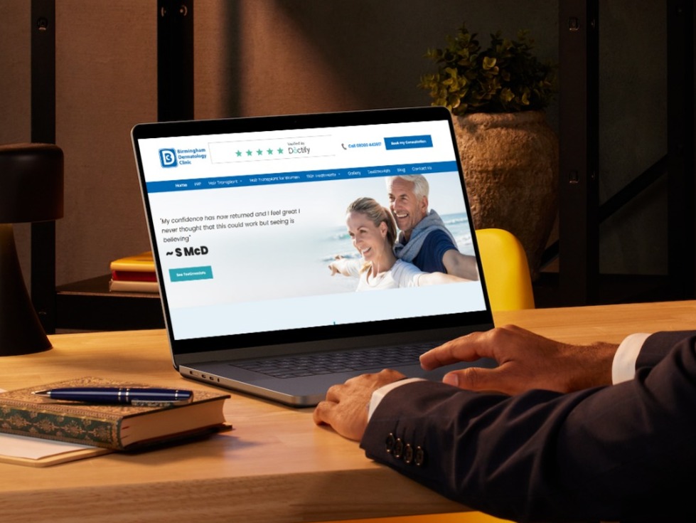 A man sat at a table, looking at his laptop screen which displays Birmingham Dermatology Clinic Healthcare website